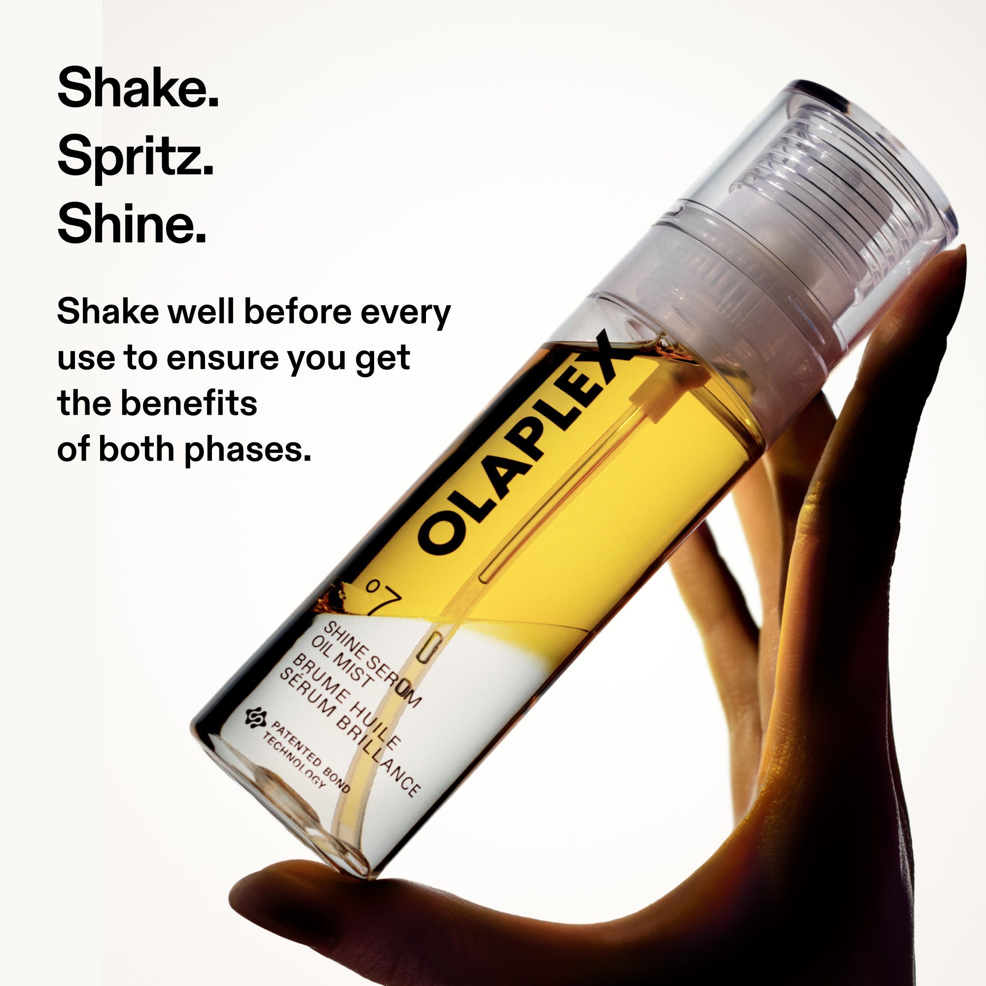 A hand silhouettes against a dark shadow, holding an angled clear bottle of Olaplex No. 7 Shine Serum Oil Mist, filled with golden liquid. To the left, white space features bold black text: "Shake. Spritz. Shine." followed by the instructional note, "Shake well before every use to ensure you get the benefits of both phases.