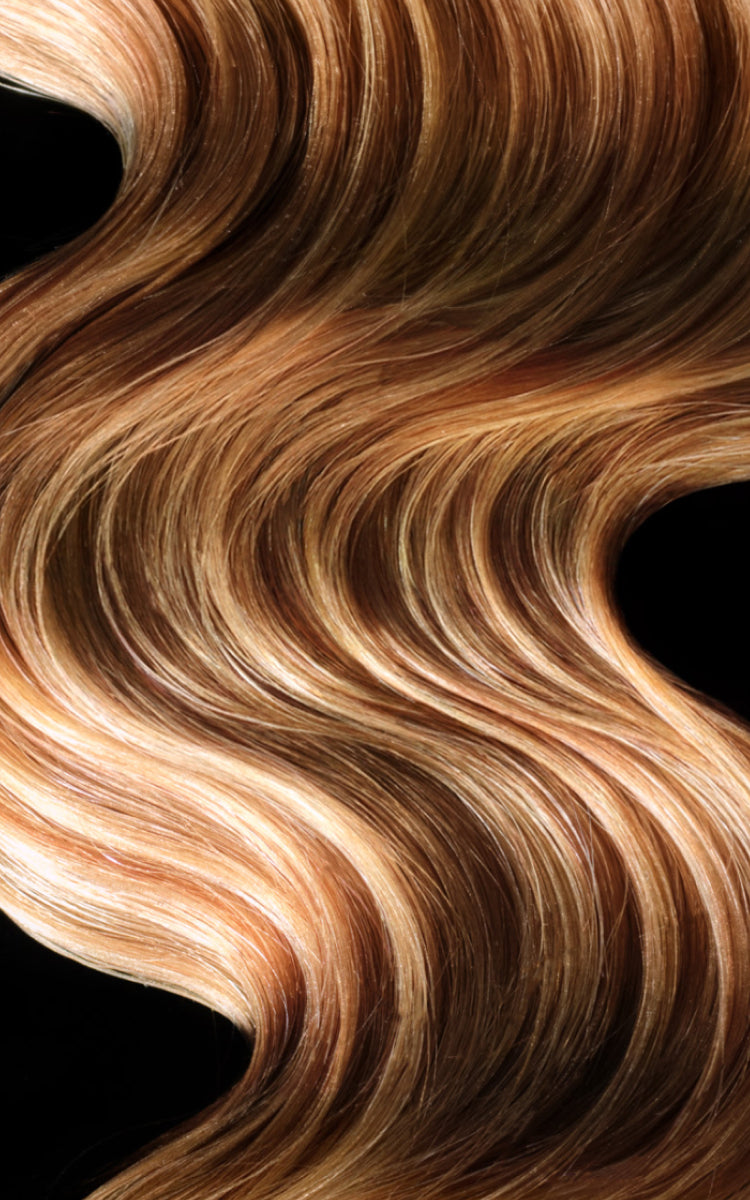 Wavy, glossy hair extensions or a wig in a warm caramel and light brown blend, shown against a solid black background.